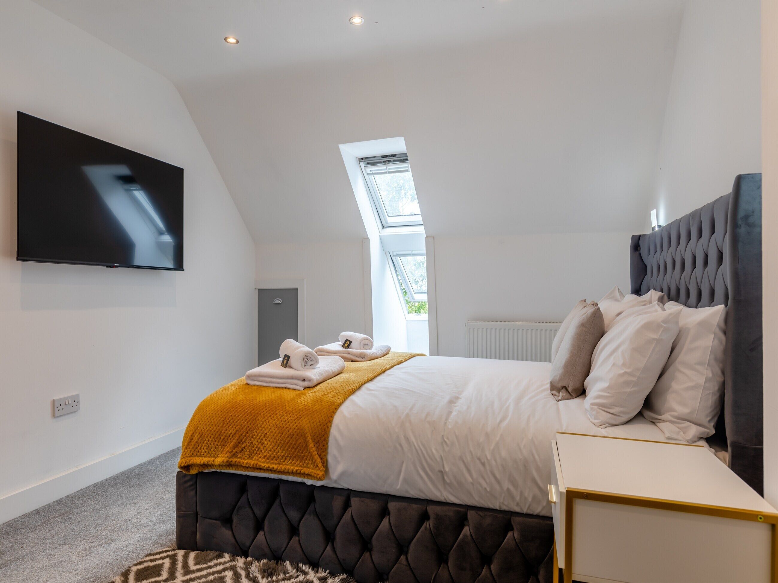 Une chambre moderne avec une tête de lit capitonnée, un téléviseur à écran plat fixé au mur, un lit bien fait avec du linge de lit blanc et un plaid de couleur moutarde, et une lucarne au-dessus du lit.