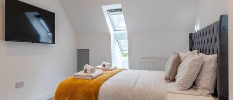 Une chambre moderne avec une tête de lit capitonnée, un téléviseur à écran plat fixé au mur, un lit bien fait avec du linge de lit blanc et un plaid de couleur moutarde, et une lucarne au-dessus du lit.