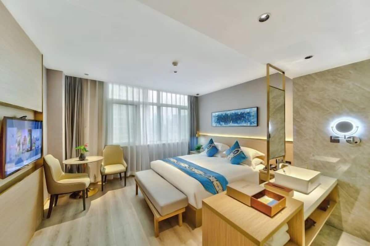 A modern hotel room with a large bed, a desk, a chair, a TV, and a bathroom with a mirror and sink.