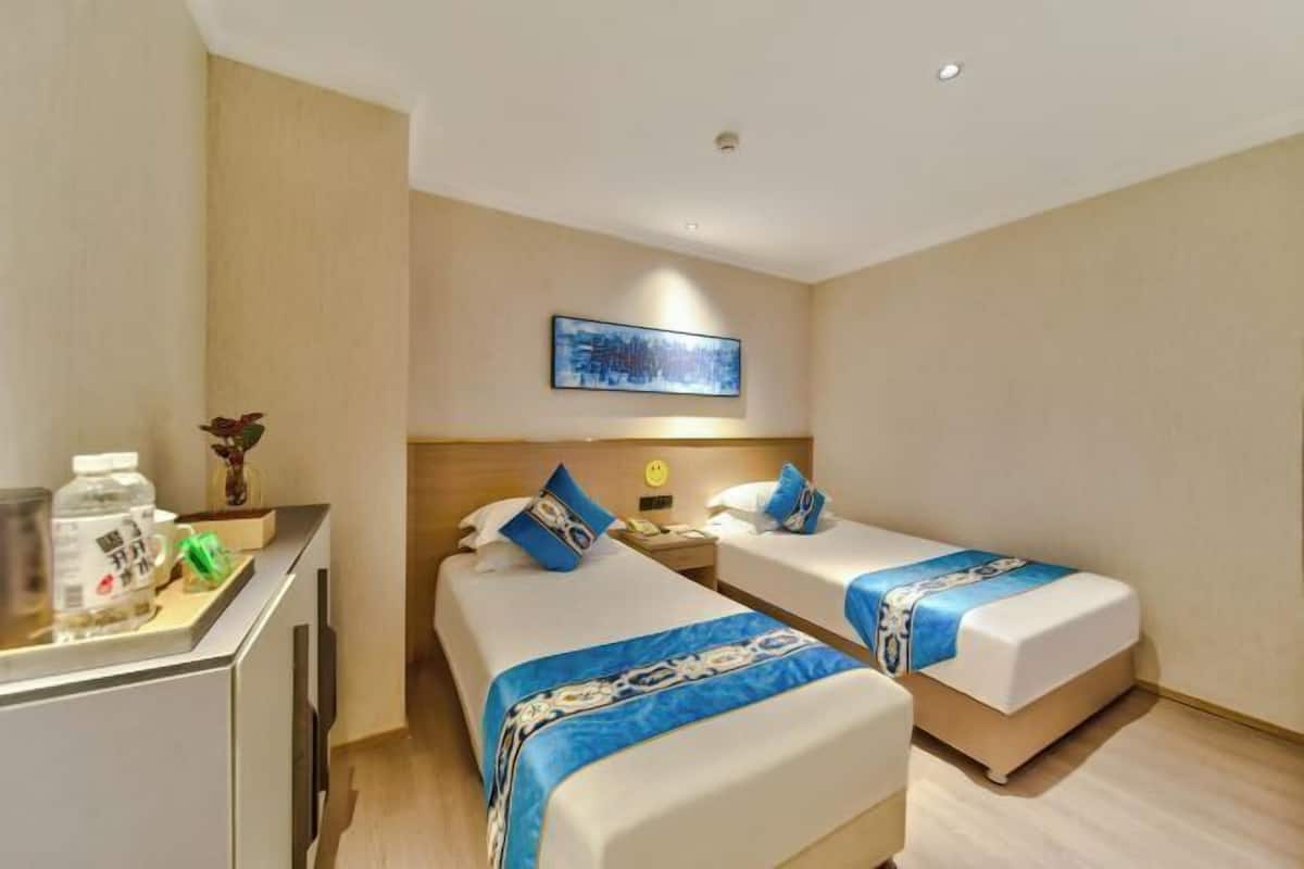 A hotel room with two beds, a desk, a nightstand, a wall-mounted TV, and a framed picture.