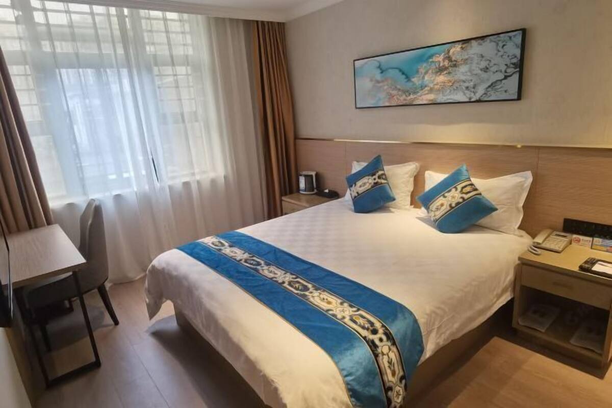 A hotel room with a large bed, a desk, and a chair. There is a window with curtains, a nightstand with a phone, and a painting on the wall.