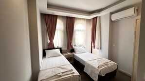 Quarto de hotel com duas camas de solteiro, mesa de cabeceira com luminária, janela com cortinas e um aparelho de ar condicionado na parede.