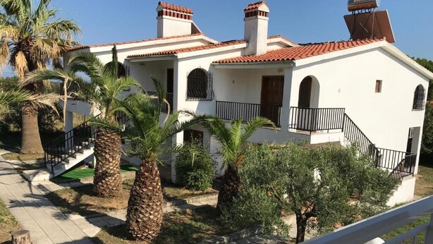 A two-story building with a white facade and red-tiled roof, featuring a central staircase, balconies with black railings, and palm trees in the courtyard. - SANTA CATALINA (Polygyros)