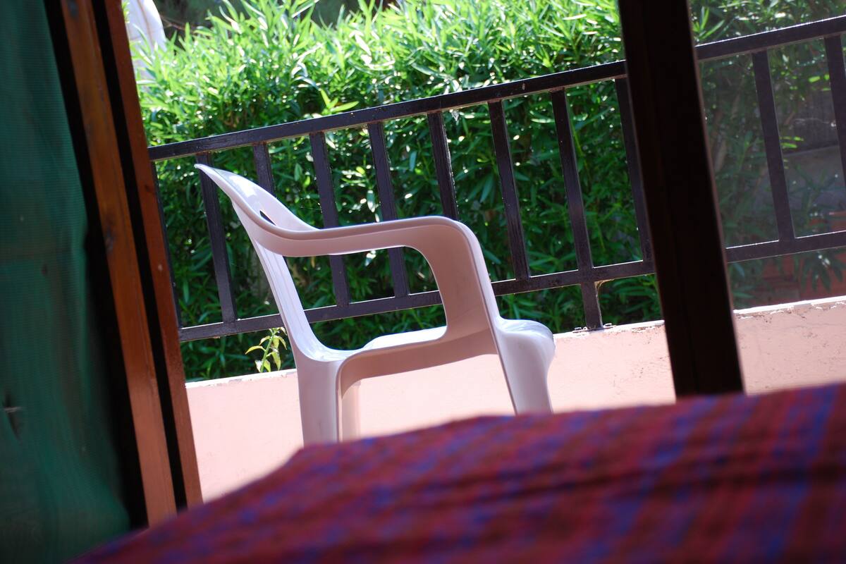 A white plastic chair in a room with a view of green foliage through a window with a black metal railing.