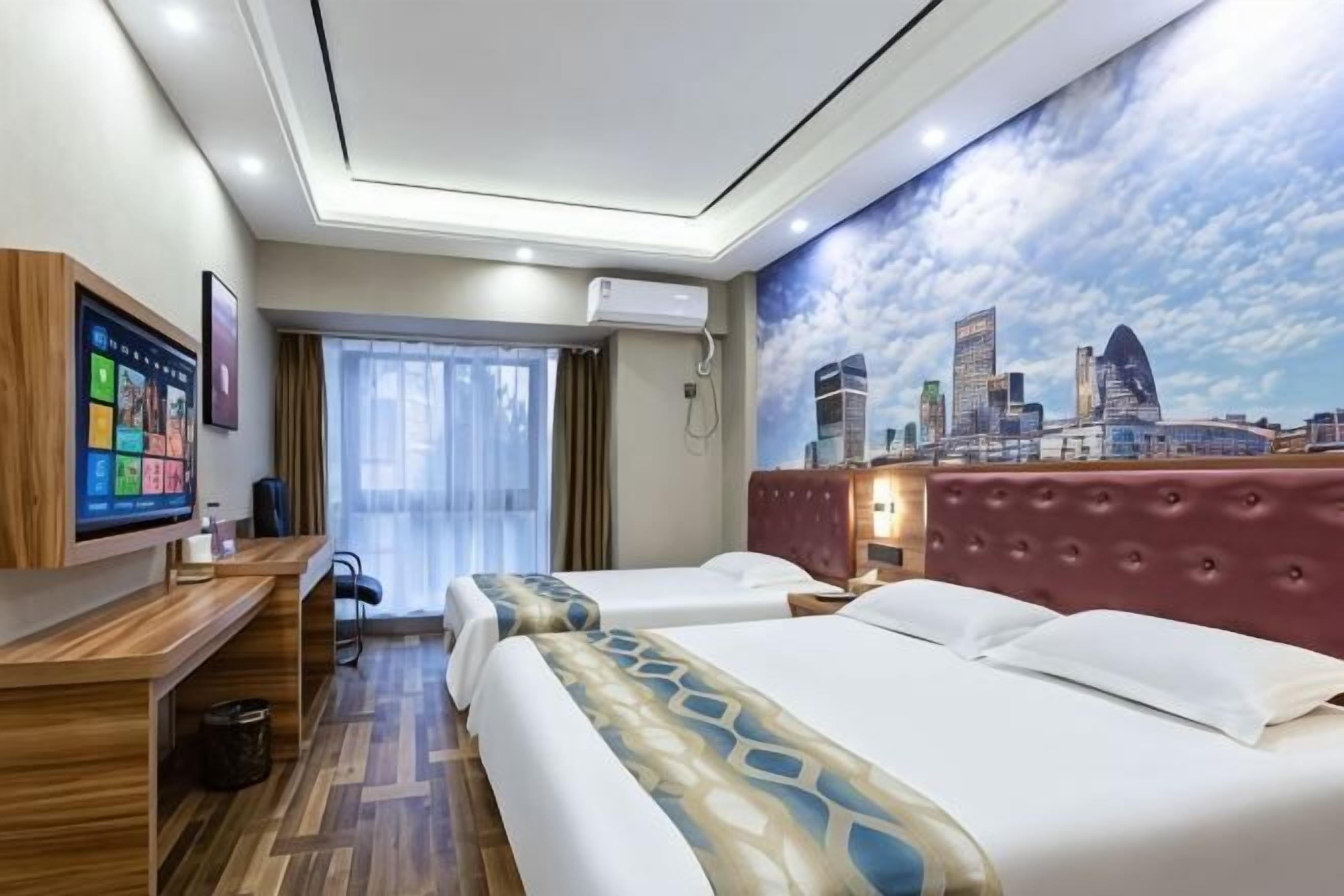 Quarto de hotel com um mural da paisagem urbana, uma escrivaninha com um computador, duas camas e uma janela com cortinas.