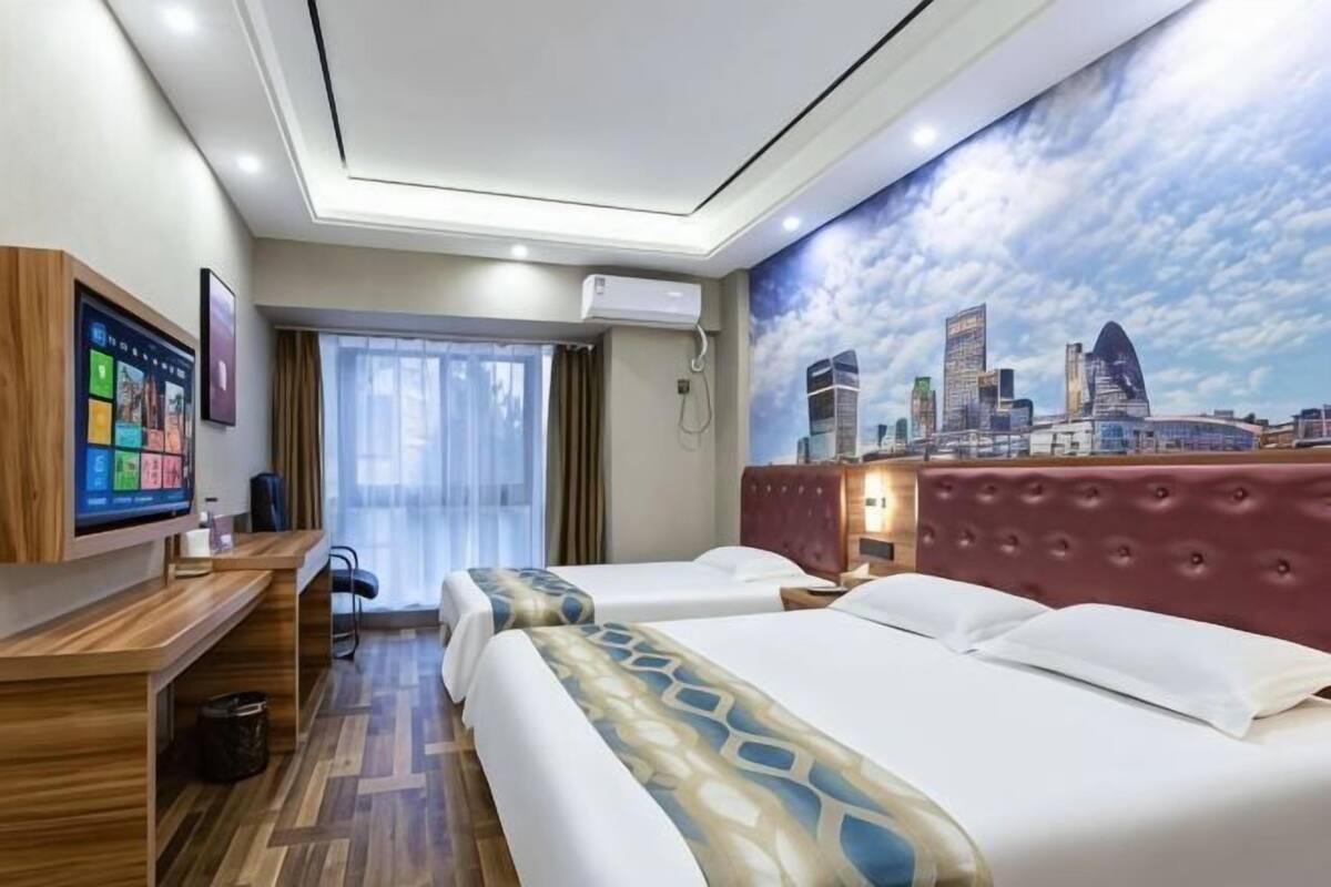 A hotel room with a cityscape mural, a desk with a computer, two beds, and a window with curtains.