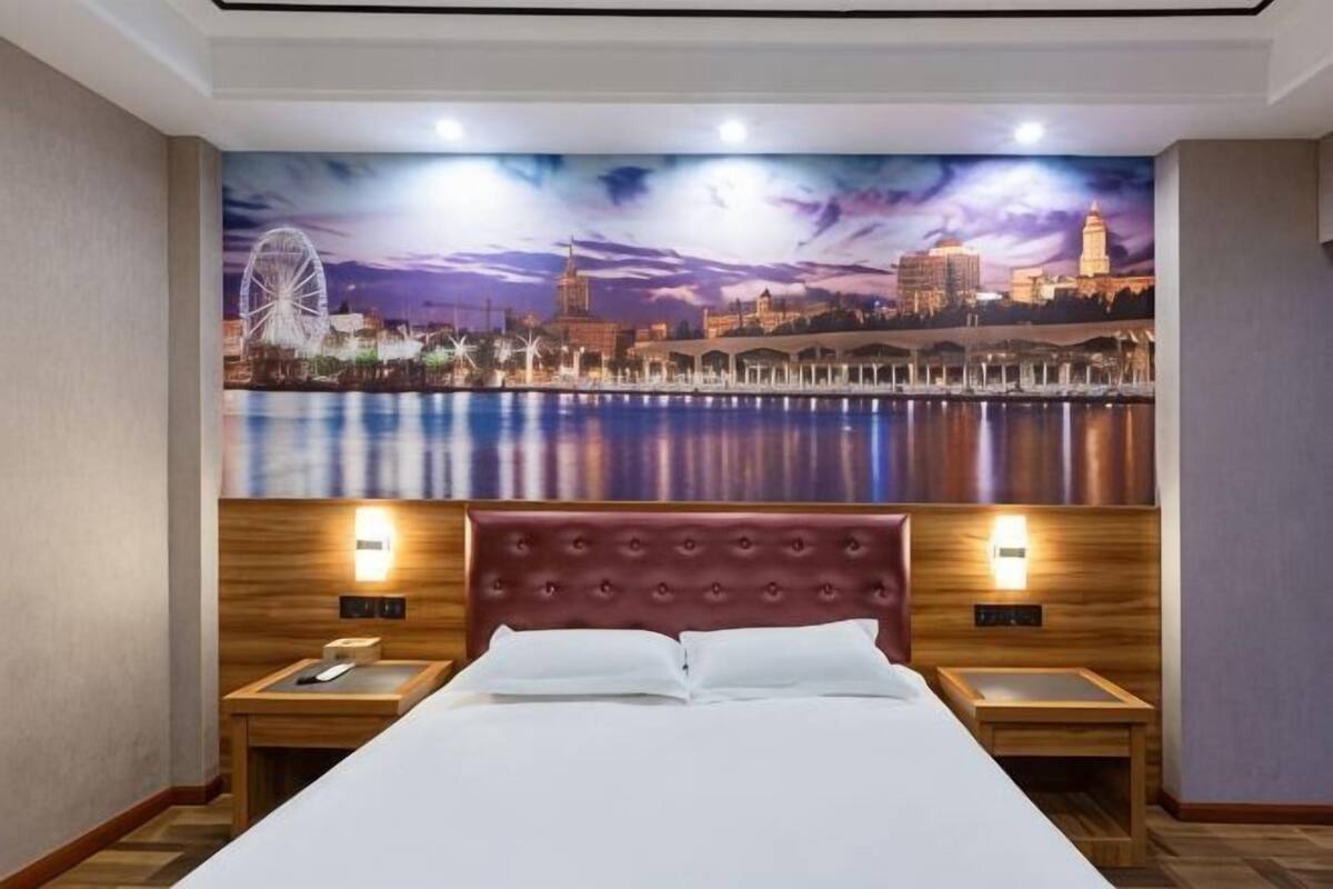 A hotel room with a large bed, wooden headboard, two bedside tables, and a cityscape mural on the wall.