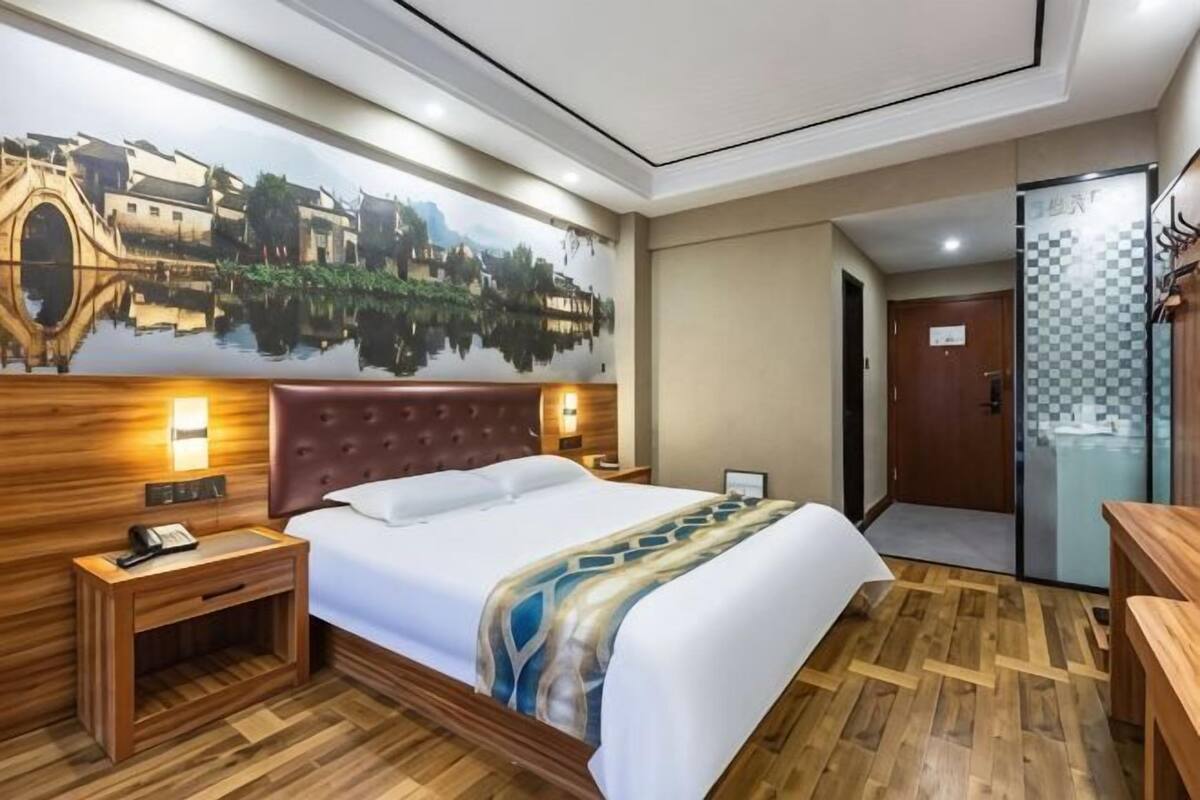 A hotel room with a bed, wooden headboard, nightstands, a wall mural, and a door to a bathroom.