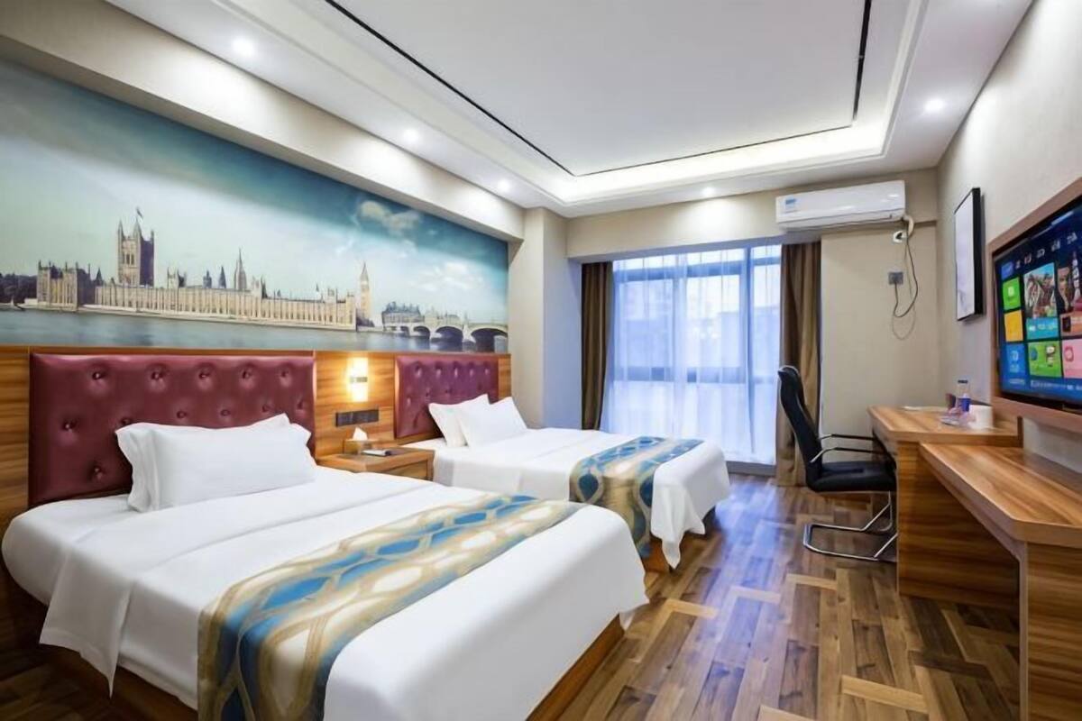 A hotel room with two beds, a desk, a chair, a TV, and a mural of a city skyline.