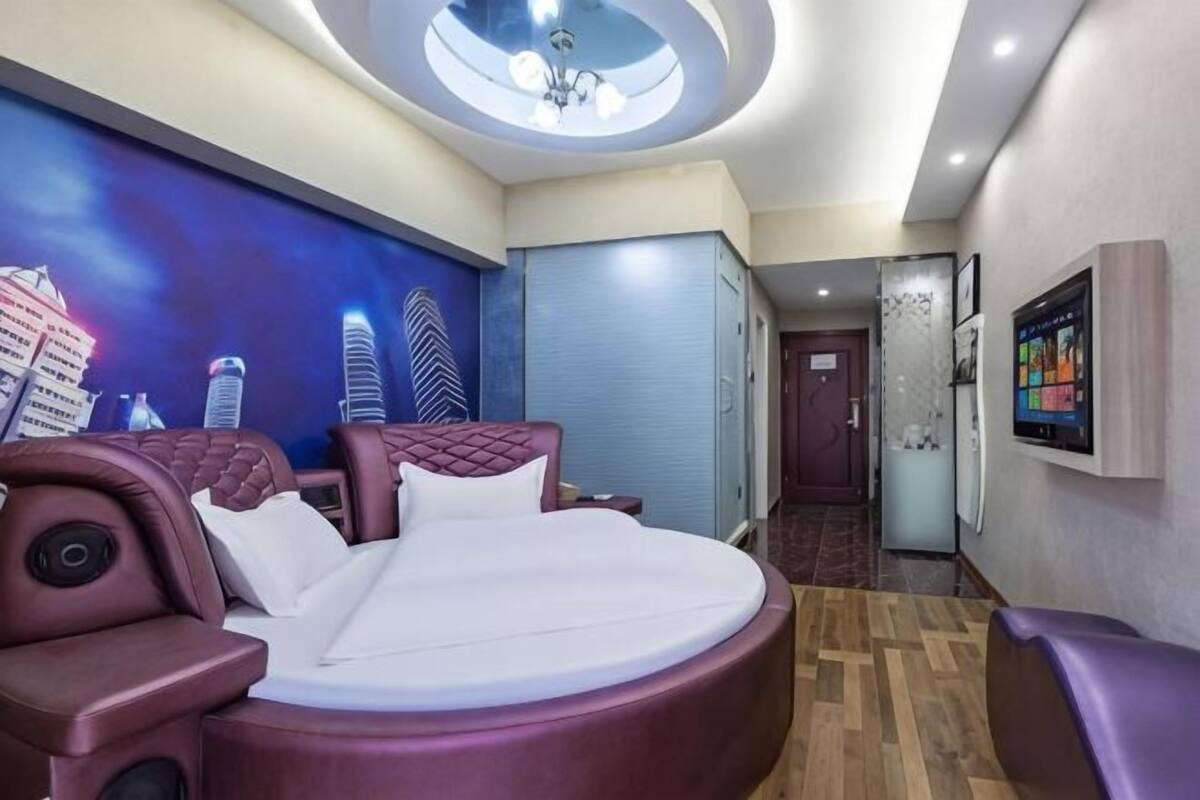 A modern hotel room with a round bed, a cityscape mural, a ceiling light, a television, and a door leading to another room.