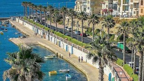 A coastal promenade with a clear blue sea, palm trees, and a row of buildings.