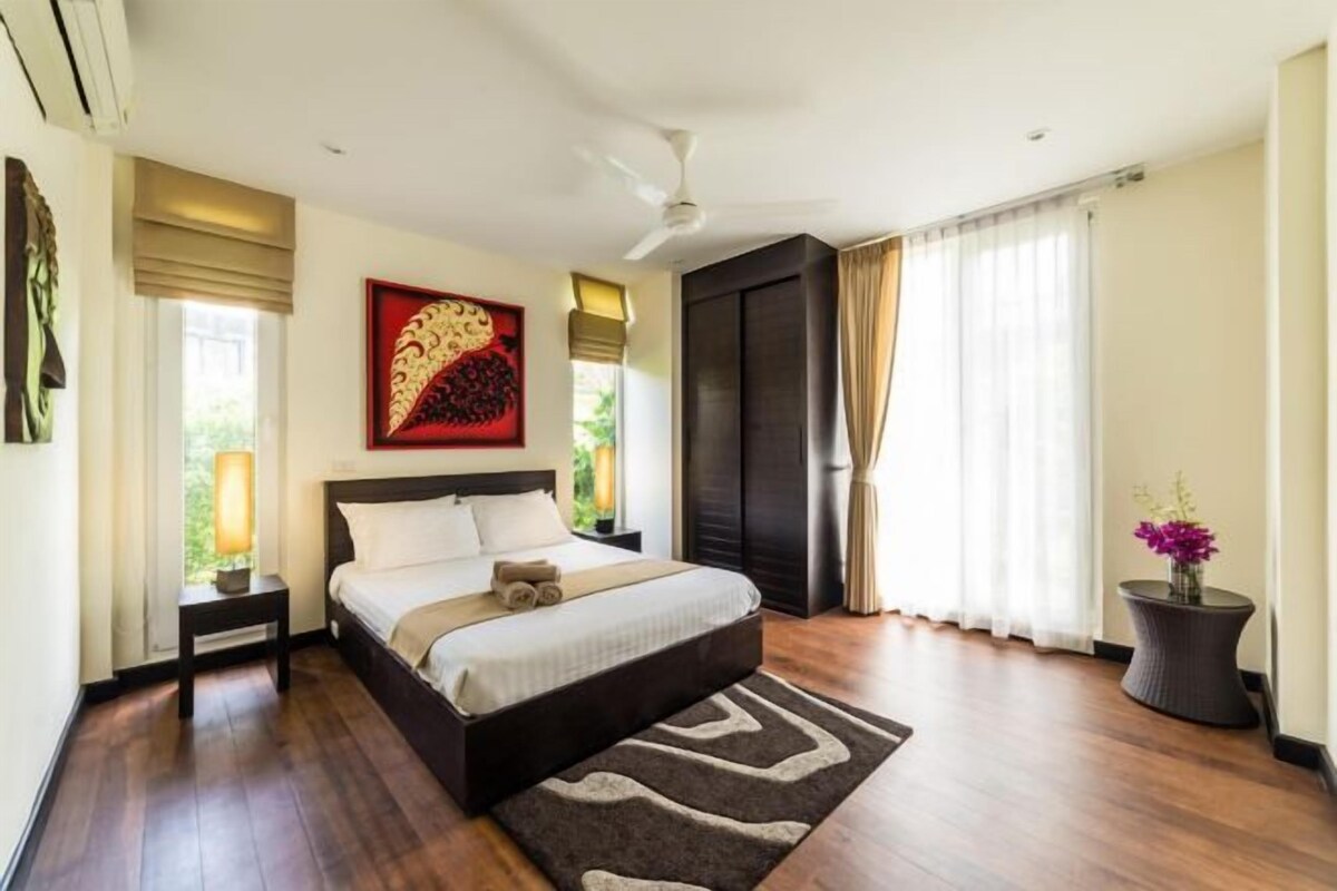 A bedroom with a bed, bedside tables, a nightstand, a window with curtains, a ceiling fan, and a potted plant.