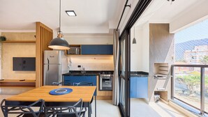 A modern kitchen with blue cabinets, a wooden dining table, and a large window overlooking a balcony. - Matira Beach 114 (Ubatuba)