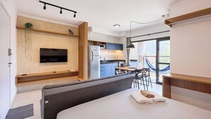 A modern hotel room with a bed, a TV mounted on the wall, a kitchen area, and a large window with a hammock outside. - Matira Beach 114 (Ubatuba)