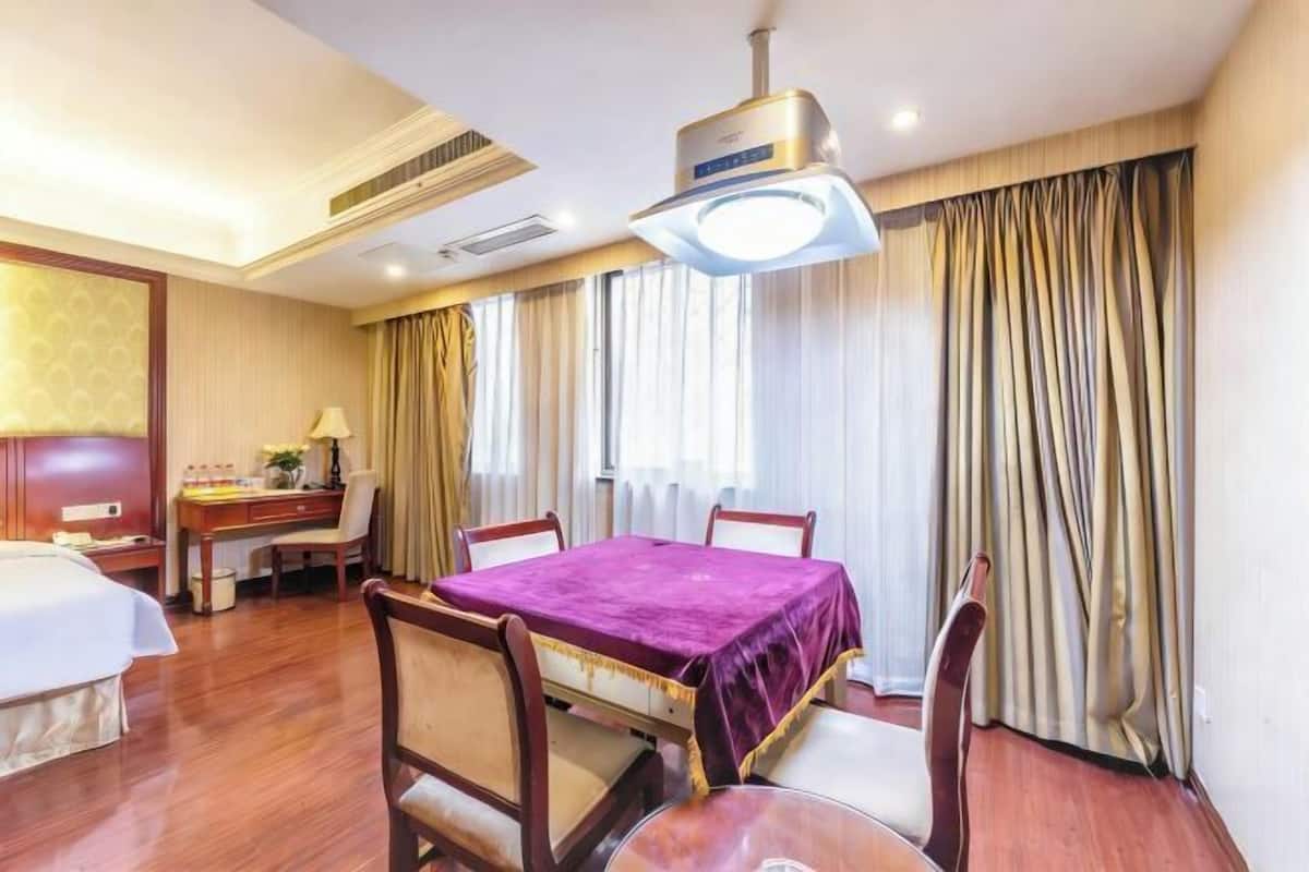 A hotel room with a bed, a desk, a dining table with chairs, a ceiling fan, and a large window with curtains.