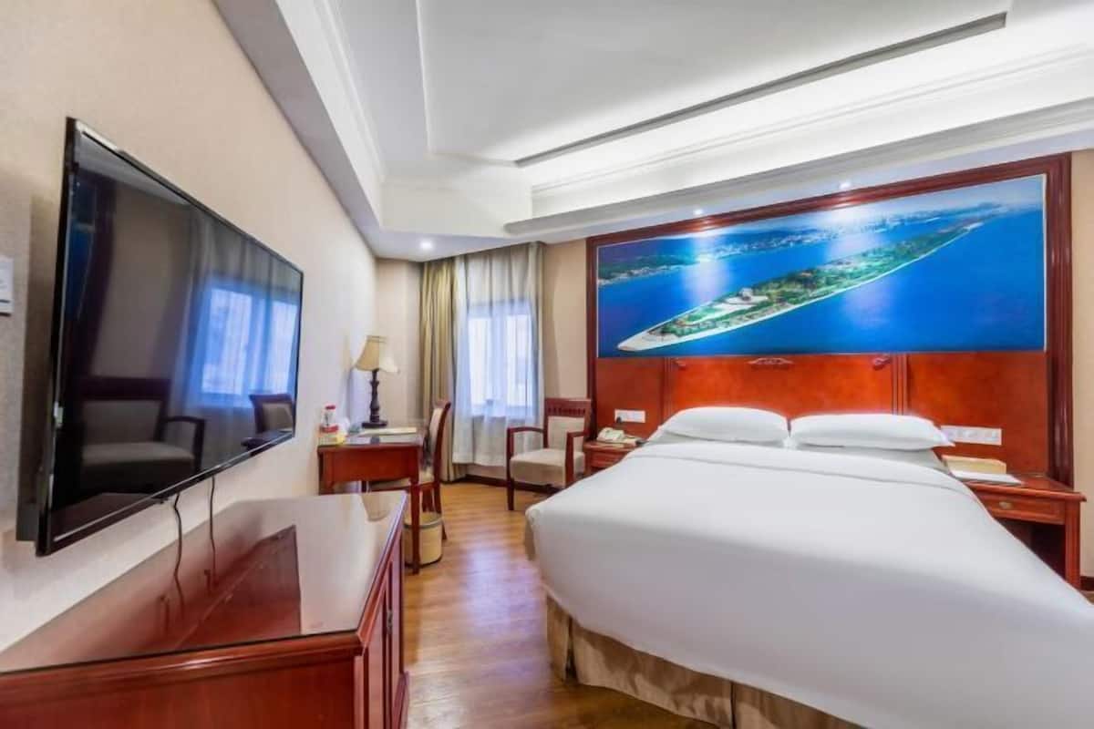 A hotel room with a bed, a TV, a desk, and a large window with curtains.