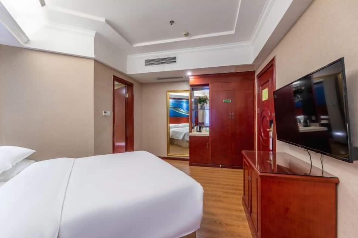 A hotel room with a bed, a TV, a wooden dresser, a mirror, and a door leading to a bathroom.
