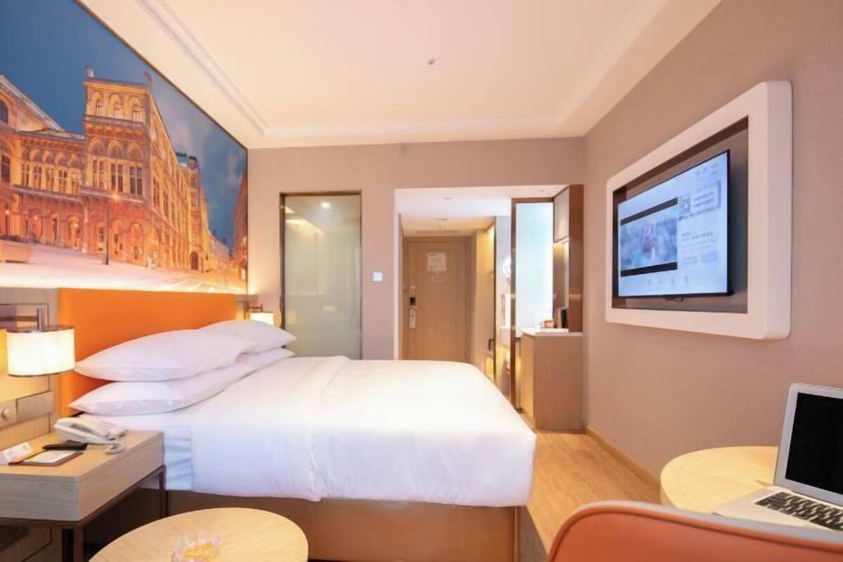A hotel room with a bed, a desk with a lamp, a laptop, a television, and a painting of a building on the wall.