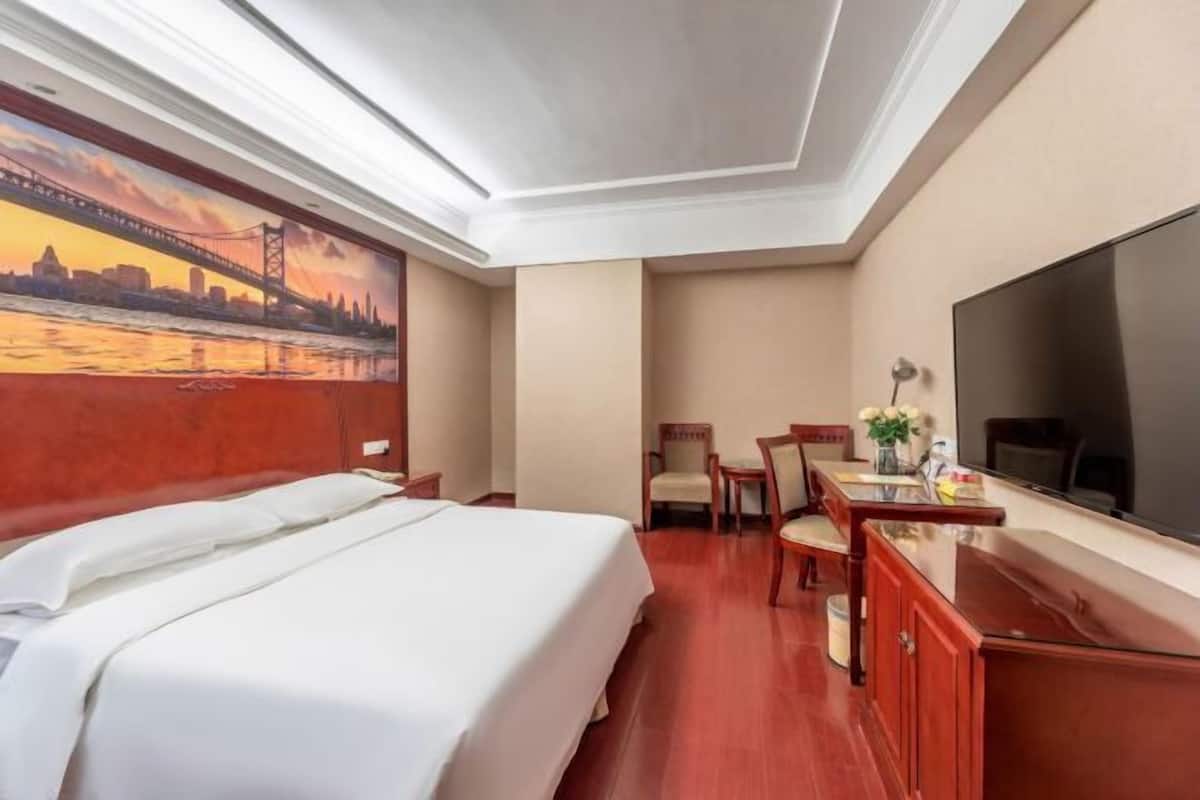 A hotel room with a bed, a desk with a chair, a TV, and a mural of a cityscape.