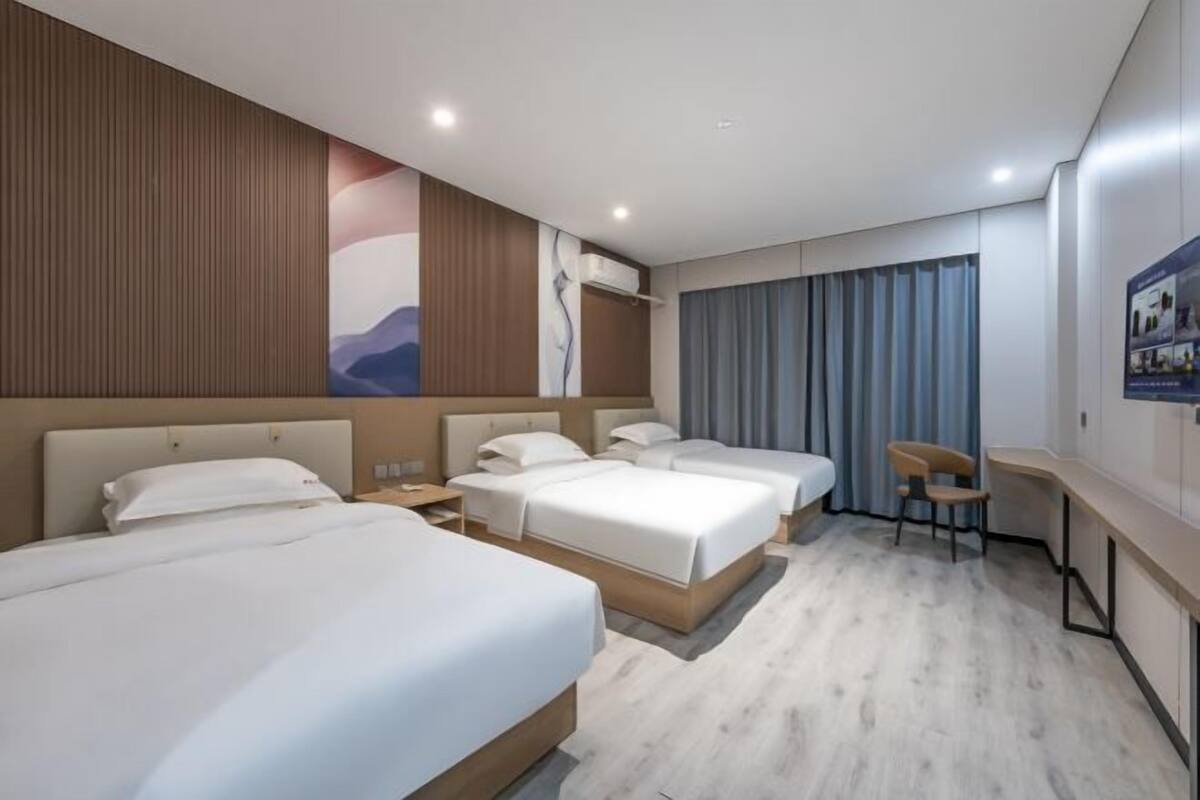 Habitación de hotel con tres camas, un escritorio, una silla y un ventanal grande con cortinas.