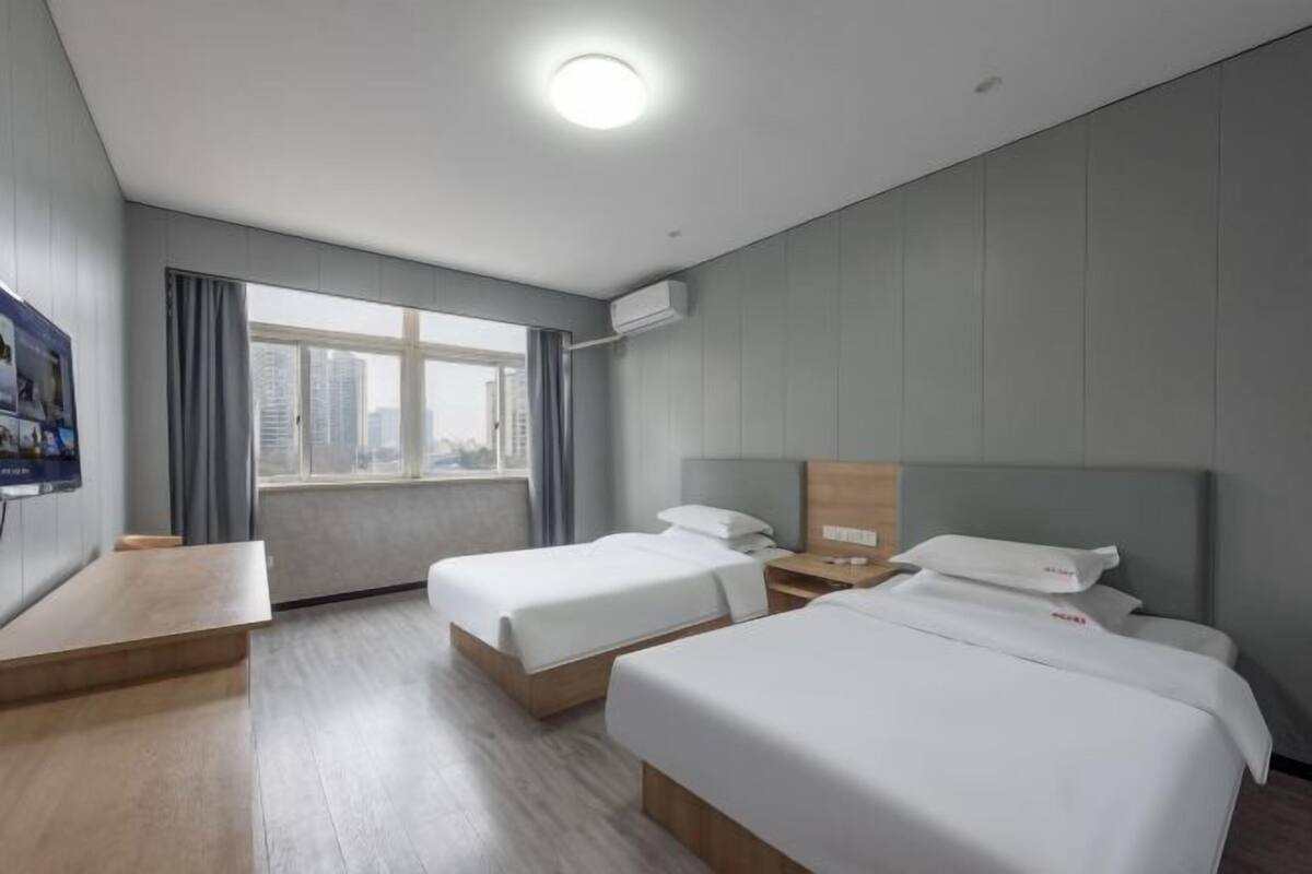 Habitación de hotel con dos camas, un escritorio de madera, un ventanal con vista a la ciudad y un equipo de aire acondicionado.