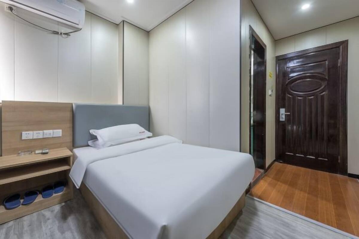 Una habitación de hotel con cama, cabecera de madera, estante con zapatos, puerta y unidad de aire acondicionado.