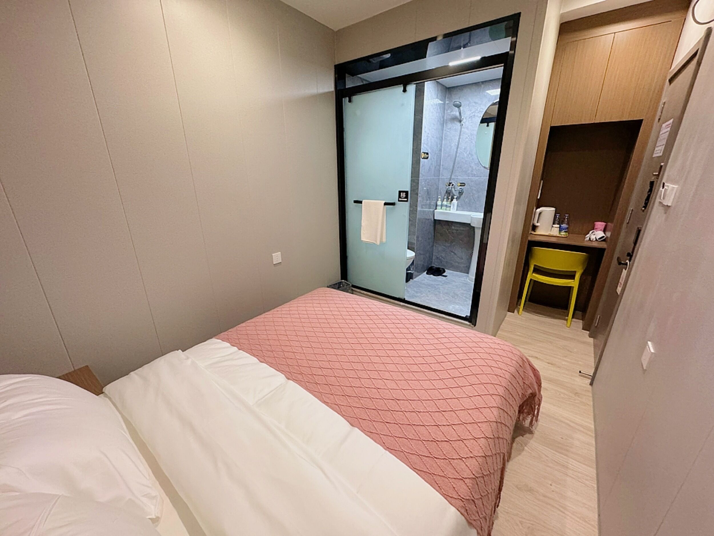 A bedroom with a bed, a small desk with a chair, and a bathroom visible through a glass door.