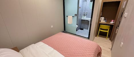 A bedroom with a bed, a small desk with a chair, and a bathroom visible through a glass door.