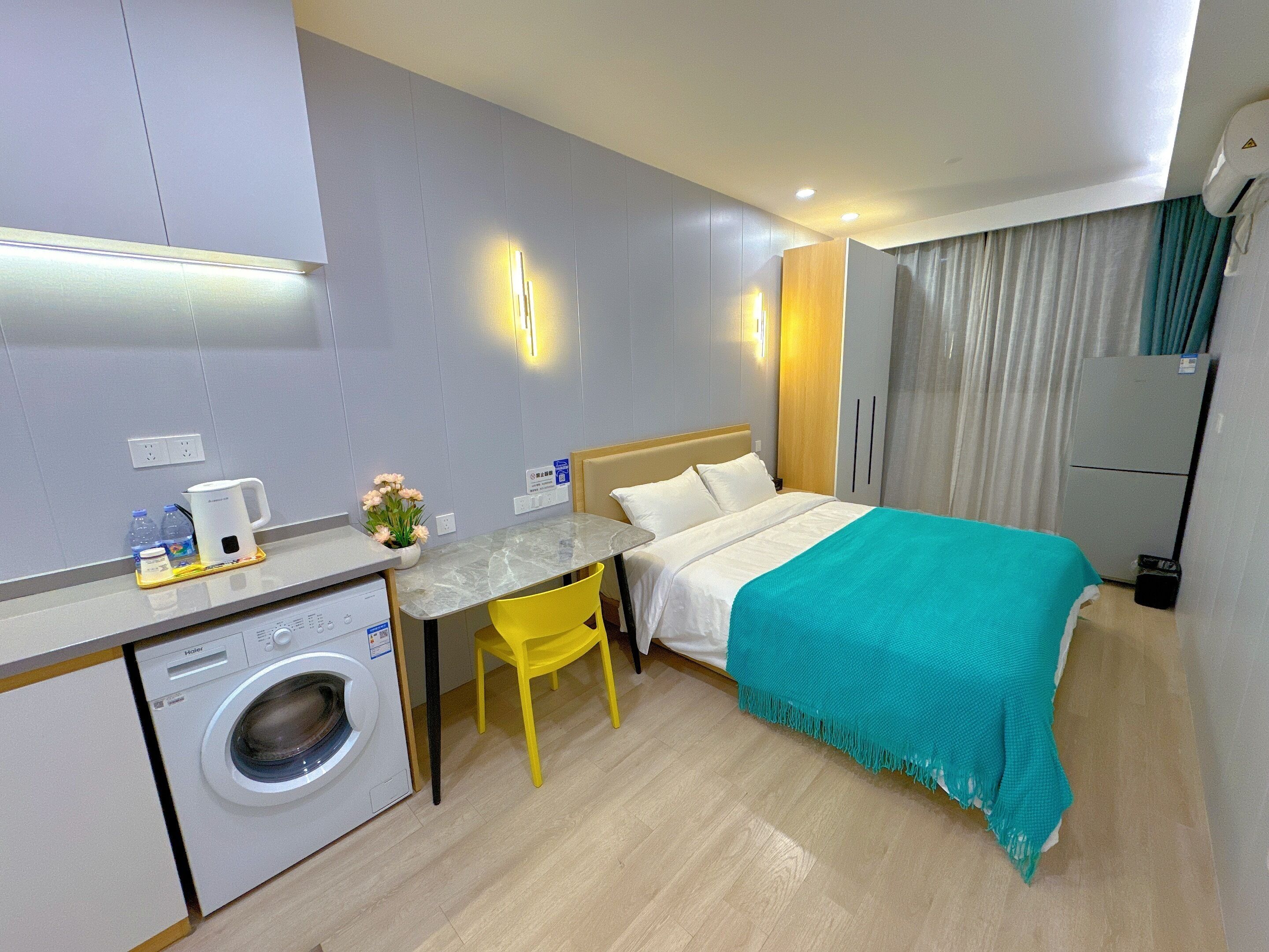 A hotel room with a bed, a desk with a chair, a washing machine, a small table with a kettle, a plant, and a refrigerator.