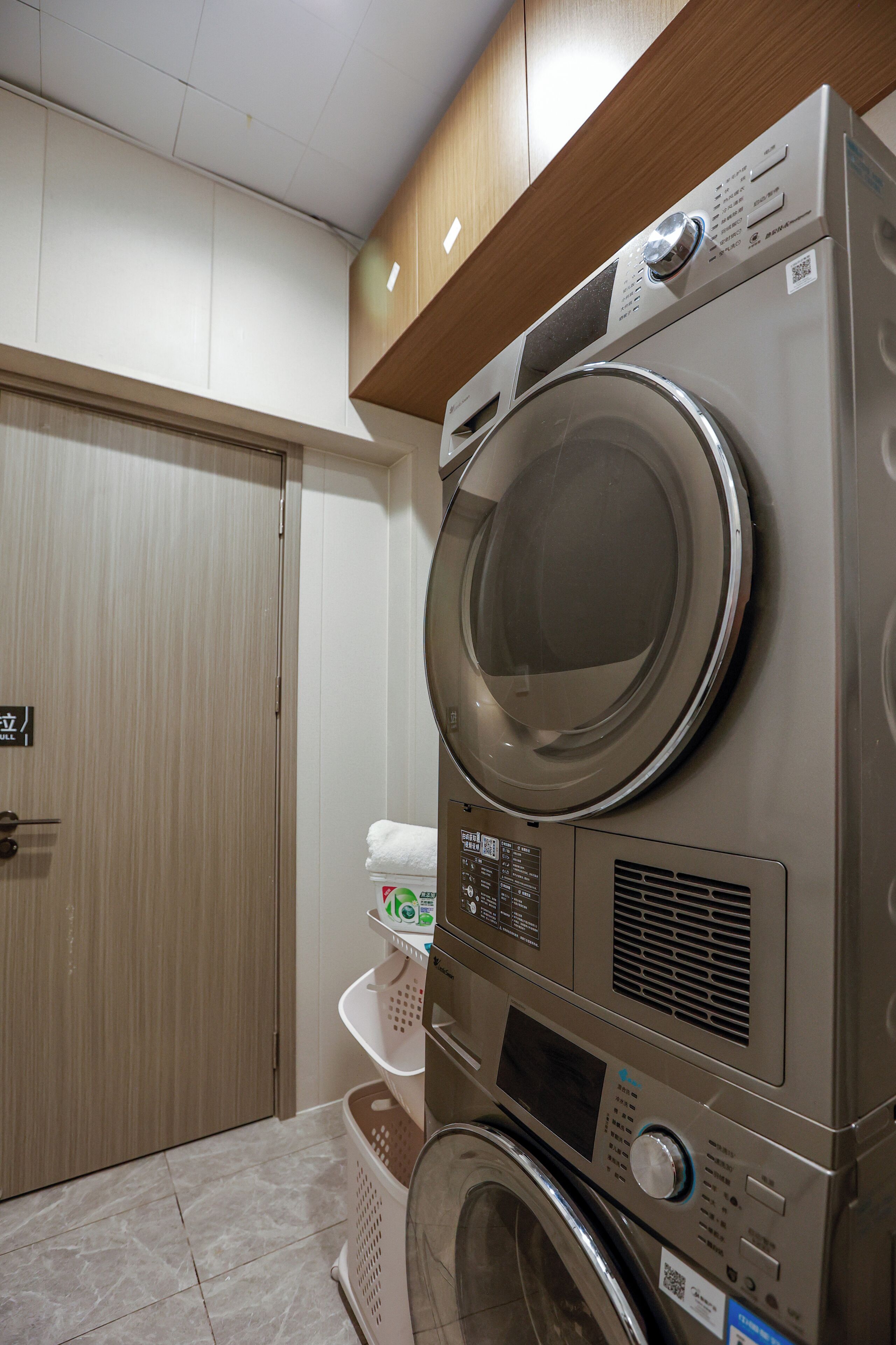 A laundry room with stacked washing machines and dryers, a wooden door, and a tiled floor.