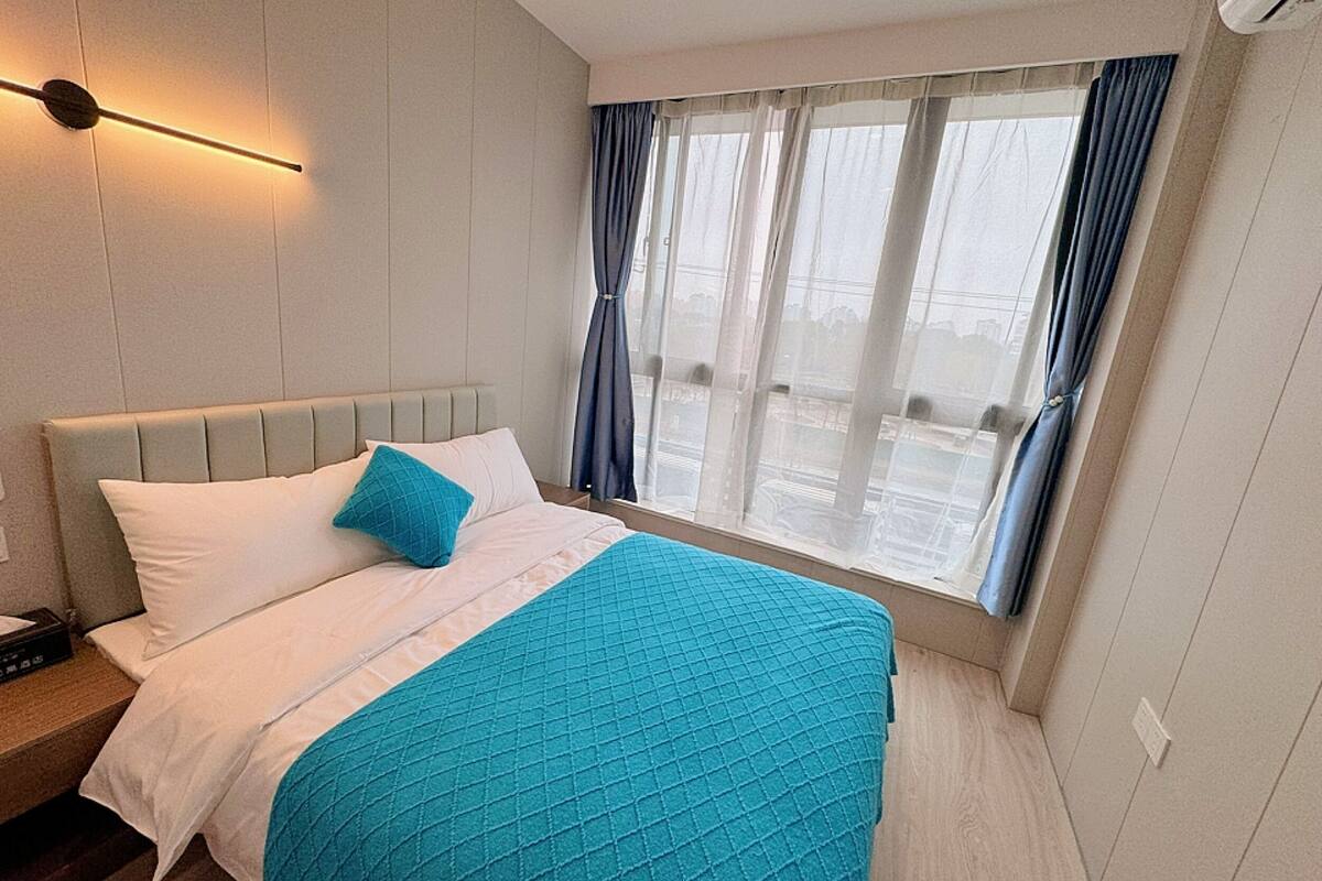 A hotel room with a bed, a nightstand, a window with curtains, and a wall-mounted light fixture.