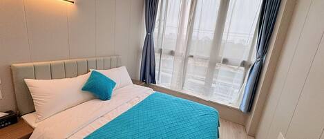 Hotellihuone, jossa on sänky, yöpöytä, ikkuna verhoilla ja seinään kiinnitetty valaisin.