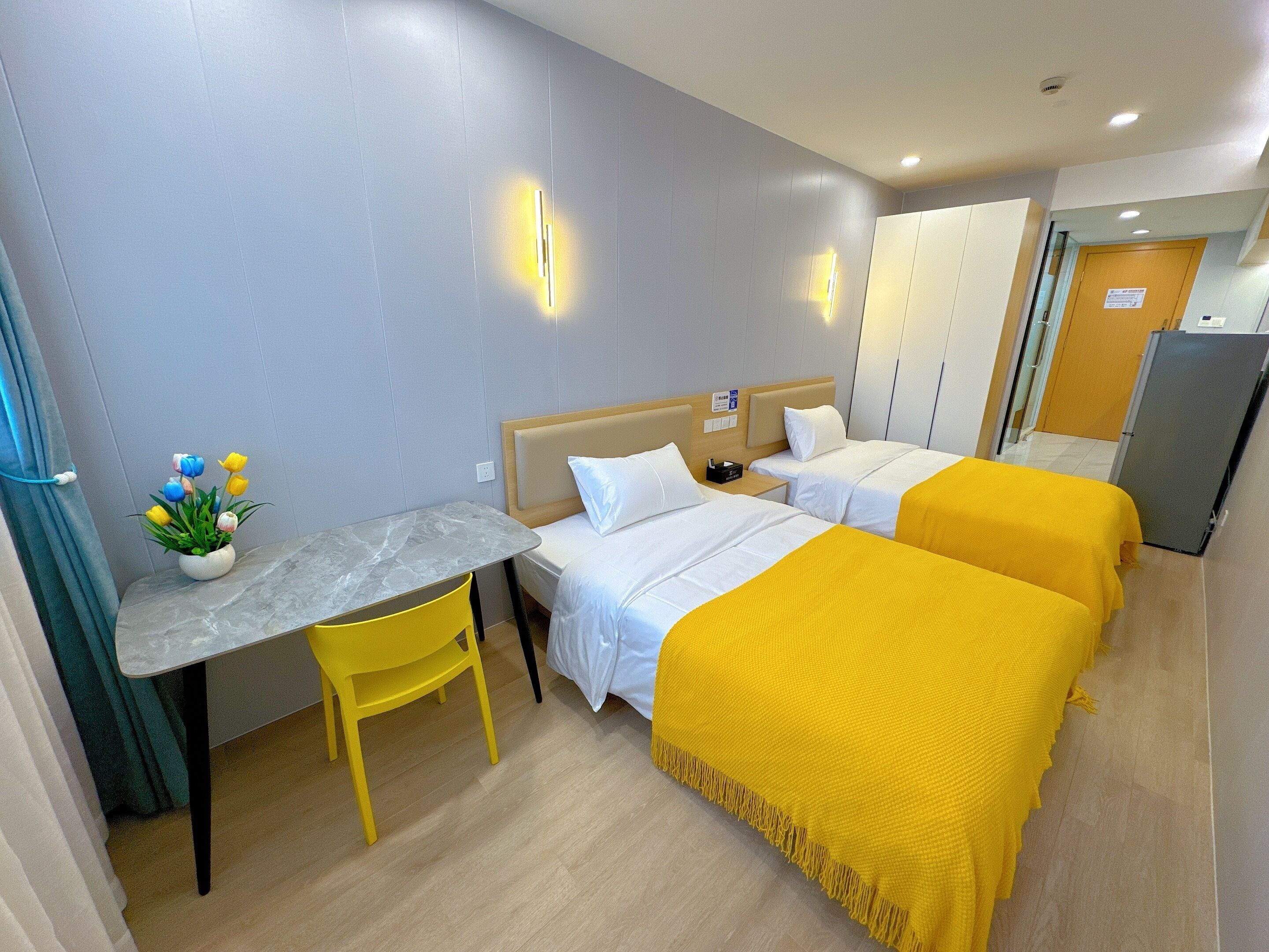 A hotel room with two beds, a small table with a chair, a plant, and a refrigerator.