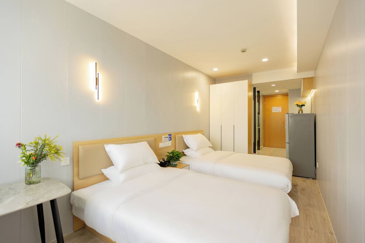 A hotel room with two beds, a small table with a vase of flowers, and a refrigerator.