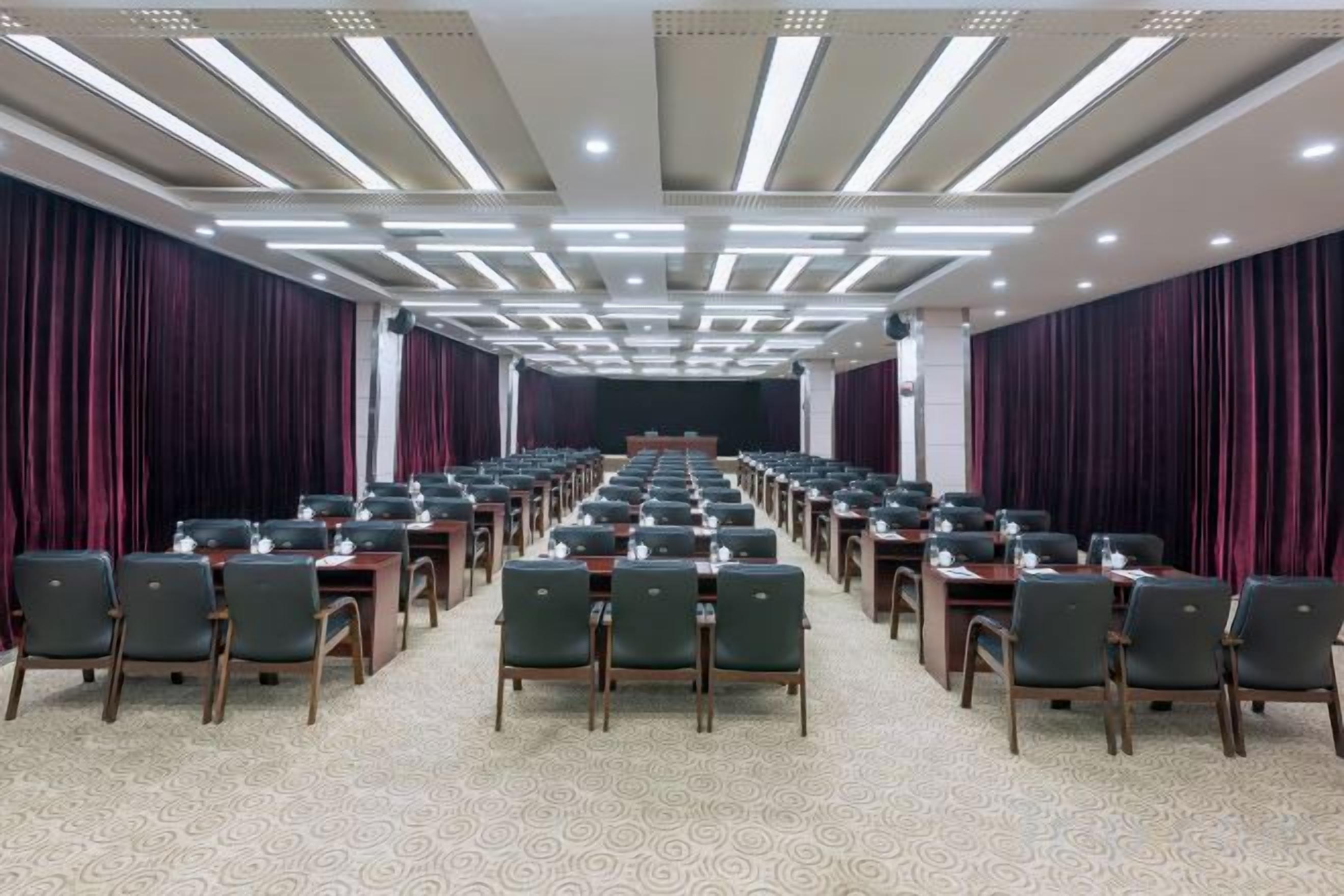 A conference room with rows of tables and chairs, a stage at the front, and a patterned carpet.