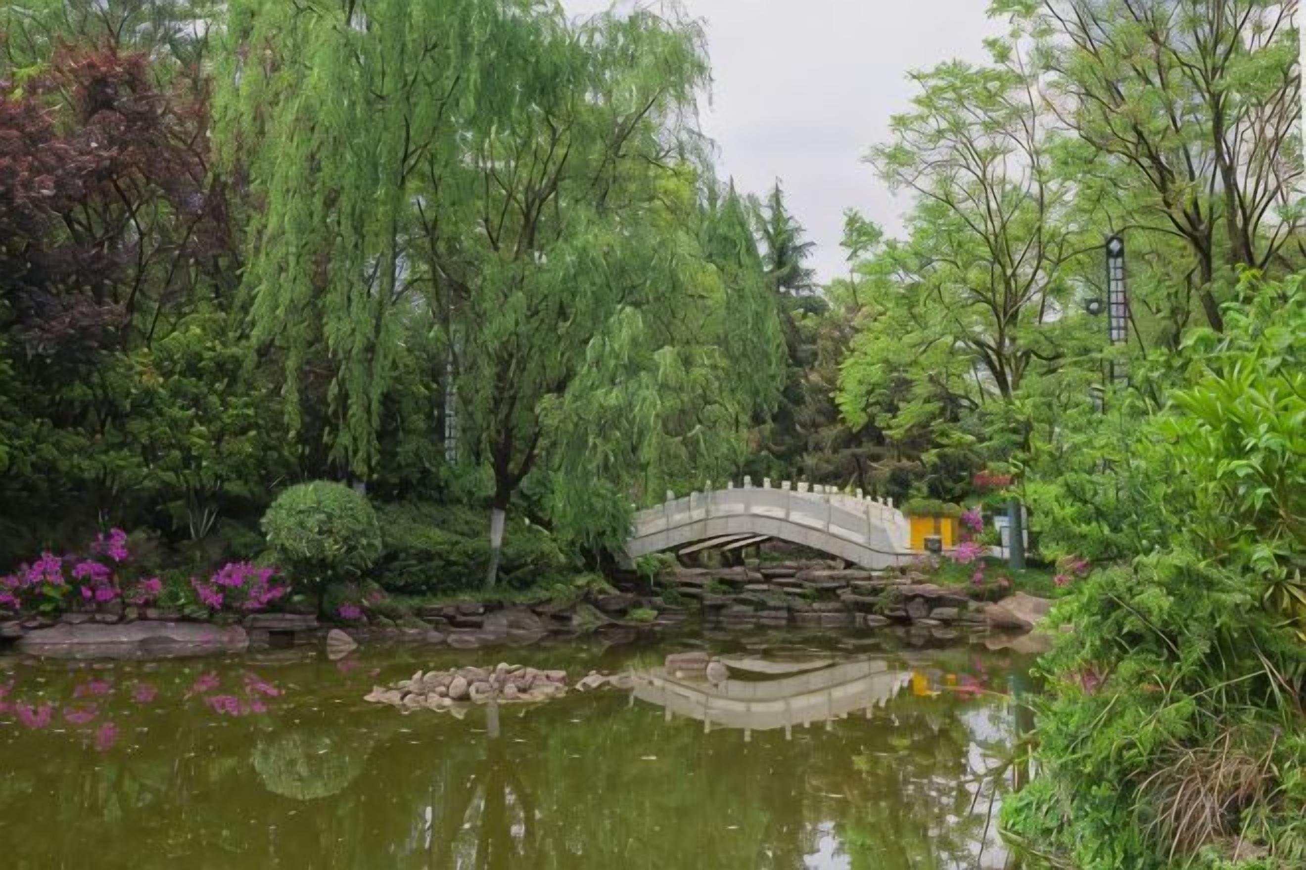 A tranquil garden with a stone bridge over a pond, surrounded by lush greenery and flowering plants.