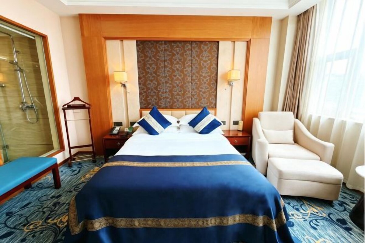 A hotel room with a large bed, a bedside table, a chair, a desk, and a shower area.