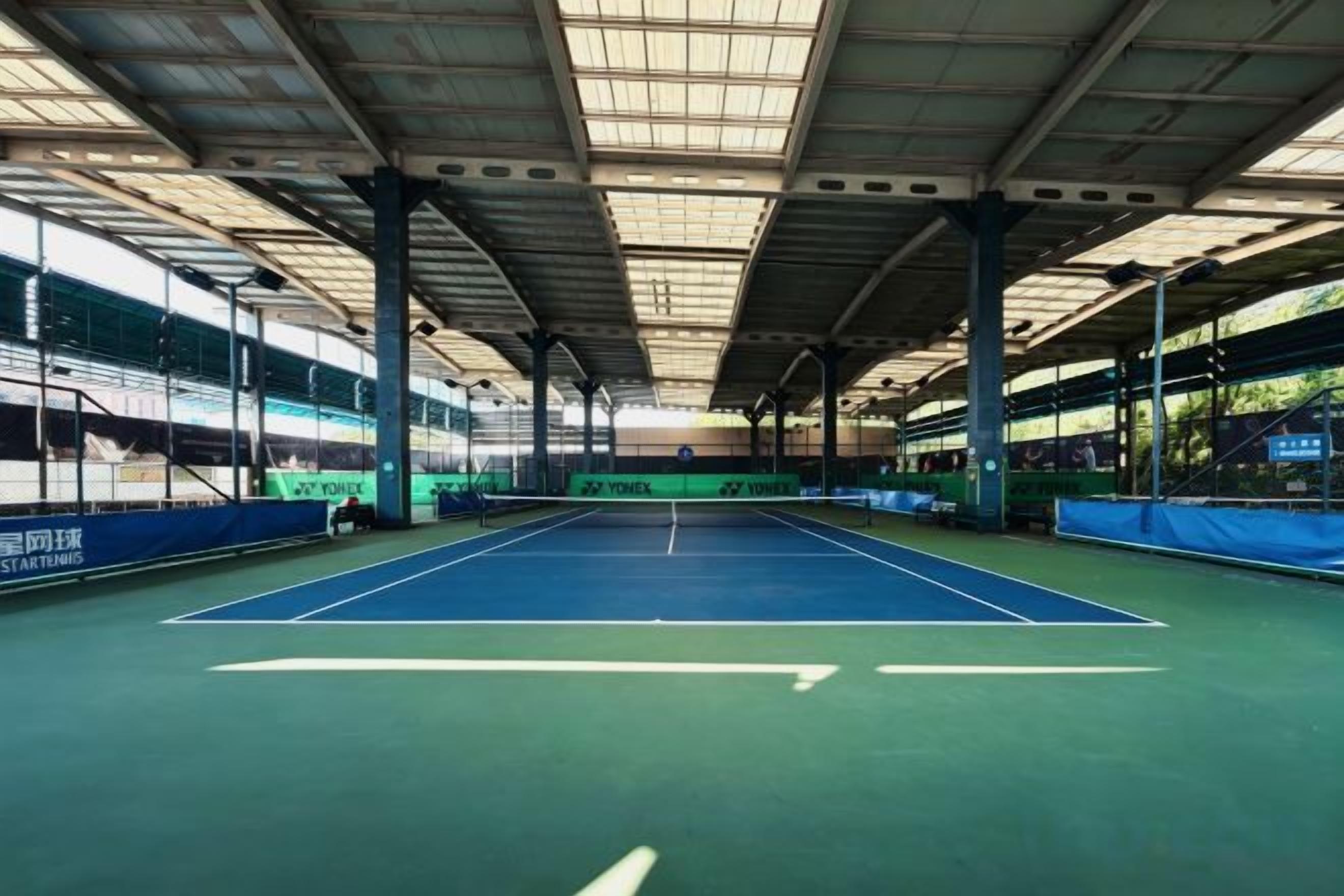 Ein Hallen-Tennisplatz mit grüner Belaga, umgeben von einer Dachkonstruktion mit sichtbaren Balken und Säulen.