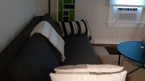 Uma sala de estar com um sofá escuro, uma luminária, uma mesinha e uma janela com persianas.