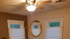 Quarto com cama, escrivaninha, cadeira, ventilador de teto e duas janelas com persianas.