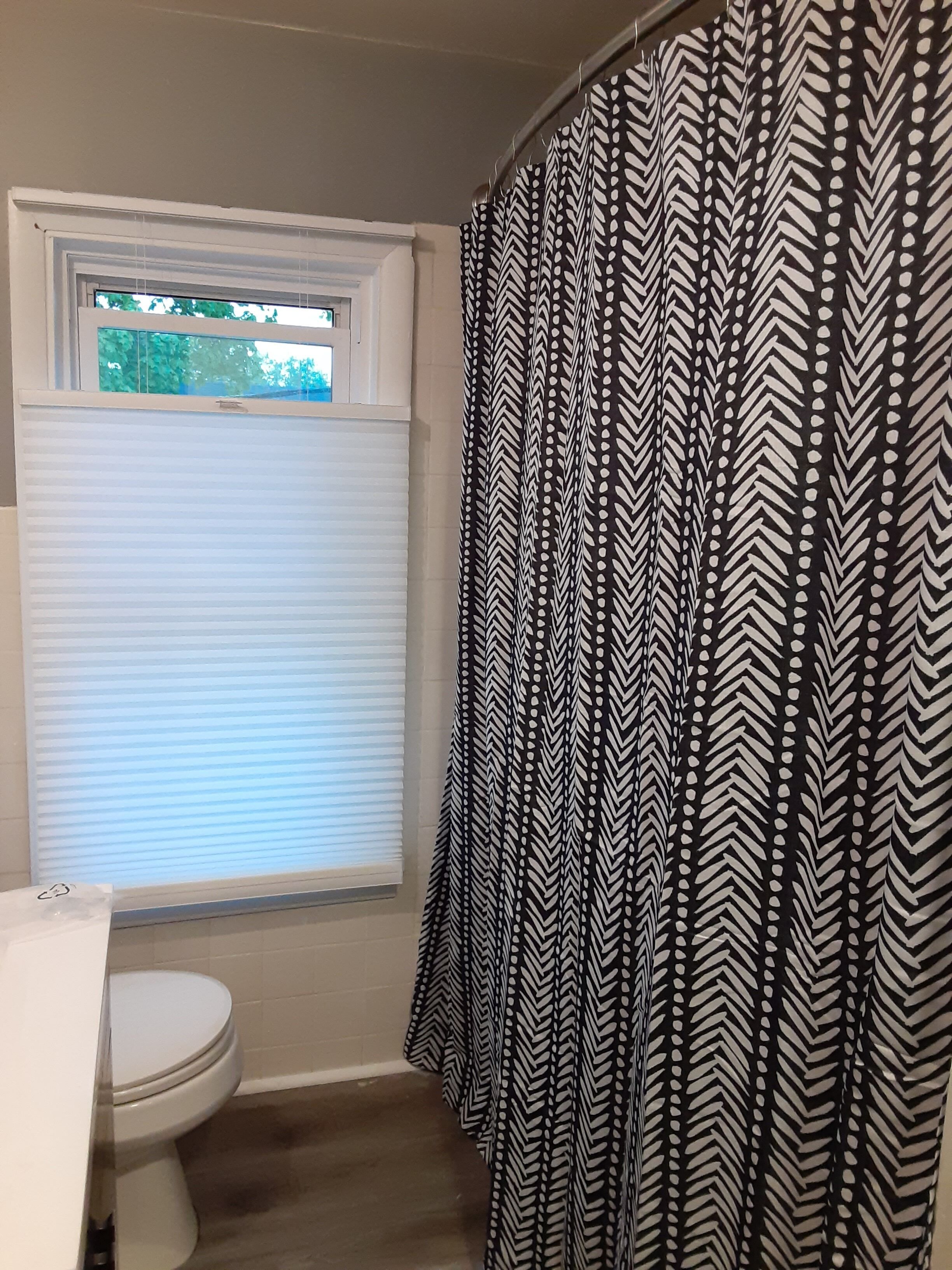 Un baño con inodoro blanco, una ventana con persianas y una cortina de ducha de estampado blanco y negro.