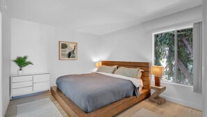 Ein modernes Schlafzimmer mit einem hölzernen Bett, weißen Wänden, einem kleinen Nachttisch mit Lampe, einer Topfpflanze und einem Fenster mit Blick auf Bäume.