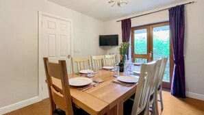 A dining room with a wooden table set for four, white chairs, a chandelier, and a window with purple curtains. - 4 bedroom accommodation in Compton Martin, near Cheddar (Compton Martin, near Cheddar)