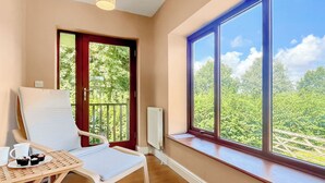 A room with a wooden floor, a white chair, a small table with a plate of food, a window with a view of greenery, and a radiator. - 4 bedroom accommodation in Compton Martin, near Cheddar (Compton Martin, near Cheddar)