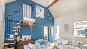 A living room with a blue wall, a staircase, a sofa set, and a window with a view. - Tyn Giat Cottage offers a perfect home for your relaxing getaways. (Amlwch and North Anglesey Heritage Coast)