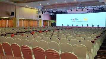 Uma sala de conferências com fileiras de cadeiras, um palco com uma tela e um pódio.