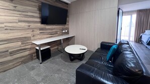 A modern hotel room with a wooden wall, a flat-screen TV, a sofa, a small round table, and a bed with a view to the outside. - One Bedroom Deluxe Spa Apartment (Hamilton)