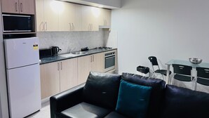 Eine kompakte Küche mit Mikrowelle, Herd und Spüle. Es gibt ein schwarzes Ledersofa mit einem türkisfarbenen Kissen sowie einen Glastisch mit Stühlen.