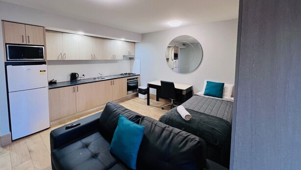 Un espace de vie compact comprenant un coin cuisine, un miroir rond, un canapé noir avec des coussins bleus et un lit avec un oreiller turquoise.