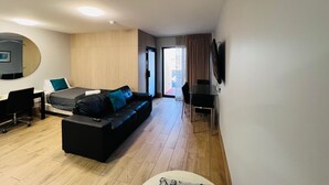 Une chambre d’hôtel moderne avec un canapé noir, un bureau avec une chaise, un téléviseur à écran plat fixé au mur et une porte-fenêtre coulissante donnant sur un balcon.